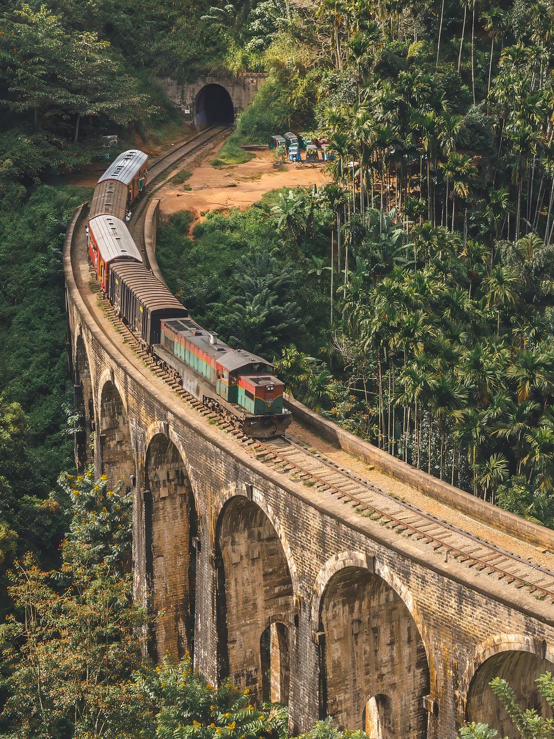 Un train circule le long du pont voûté en pierre aux neuf arches au Sri Lanka, un chef-d'œuvre d'ingénierie entouré d'une forêt verdoyante. Le train entre dans un tunnel en arrière-plan, avec un groupe de personnes debout à proximité au sol, observant la scène. Cette scène évocatrice est magnifiquement capturée dans "Un billet pour le passé" (Sri Lanka) de Fine Art Shots.