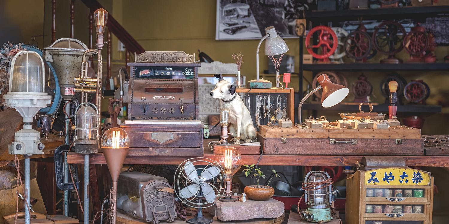 Un magasin d'antiquités encombré présente des objets vintage. La table en bois contient des lampes rétro, une vieille radio et une figurine de chien. Divers objets, dont des ventilateurs et des valises, remplissent les étagères du fond. Un éclairage chaleureux crée une ambiance nostalgique avec "La mémoire des objets" de Fine Art Shots qui ajoute au charme de l'exposition.