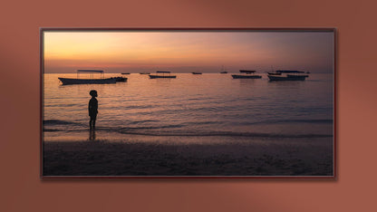 Une photographie encadrée sur "L'enfant et la plage" de Fine Art Shots (Tanzanie) représente un enfant debout sur une plage au coucher du soleil. La silhouette de l’enfant fait face à la mer calme, où sont ancrés plusieurs bateaux. Le ciel est teinté de teintes chaudes et oranges, se reflétant sur les douces vagues.