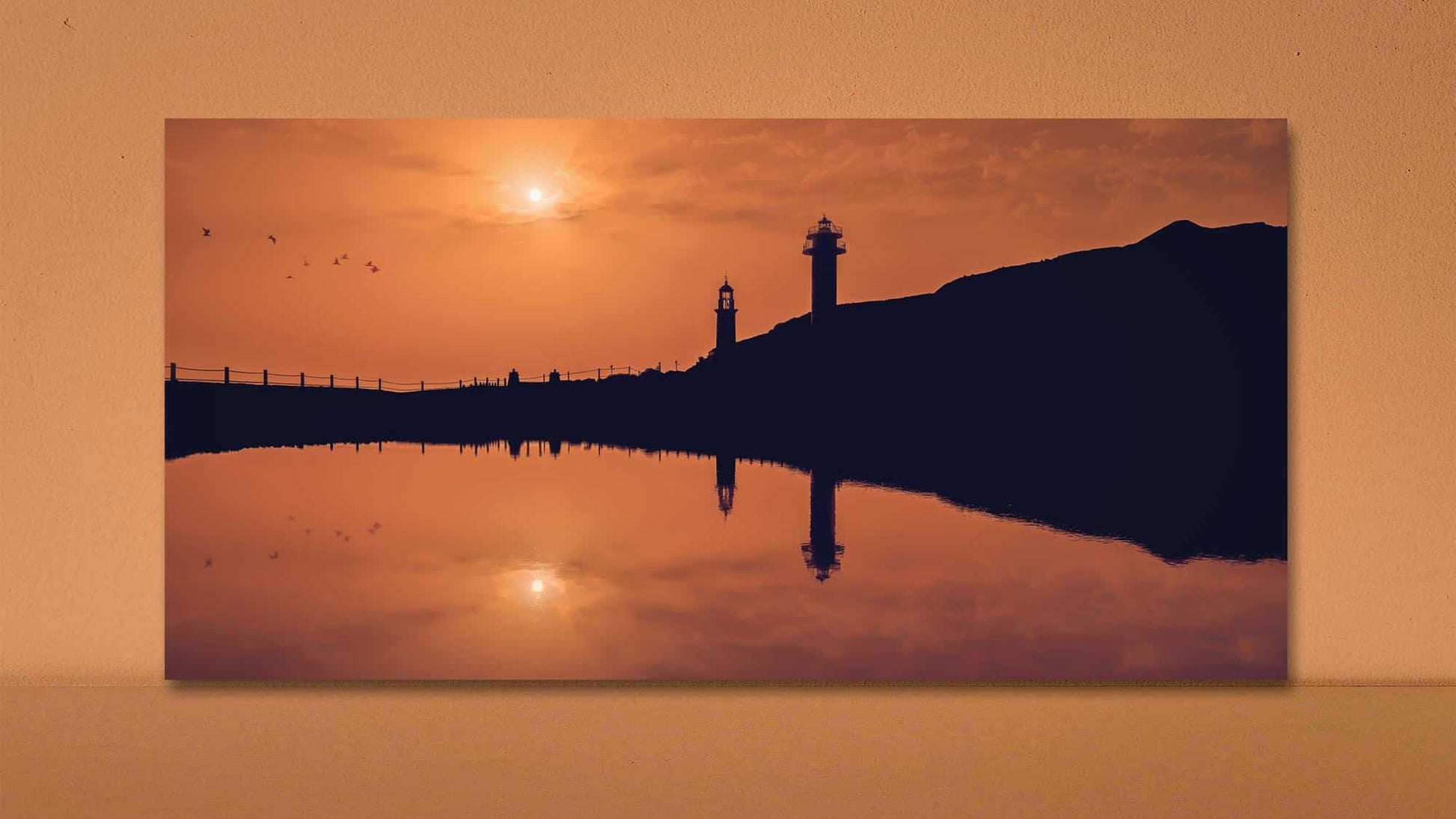 Photographie d'un coucher de soleil serein sur un plan d'eau calme, avec les silhouettes de deux phares sur le flanc d'une colline. Le ciel est d'un orange chaud et les oiseaux volent près du soleil, créant une scène tranquille et pittoresque. "Un mercredi soir sur la terre" (Îles Canaries) de Fine Art Shots est accroché au mur.