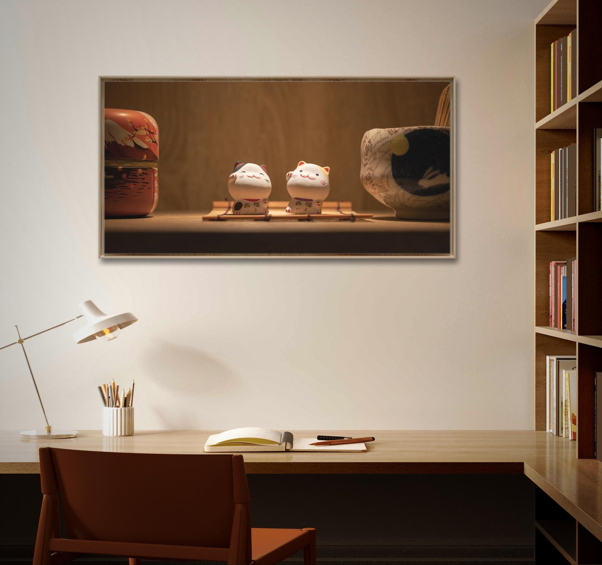 Un bureau en bois avec une chaise marron, équipé d'une lampe de bureau blanche, des crayons dans un porte-crayon et un cahier fermé. "Les chats porte-bonheur" (Taïwan) de Fine Art Shots et un bol sont exposés dans un cadre sur le mur blanc au-dessus du bureau. Une bibliothèque en bois est partiellement visible à droite.