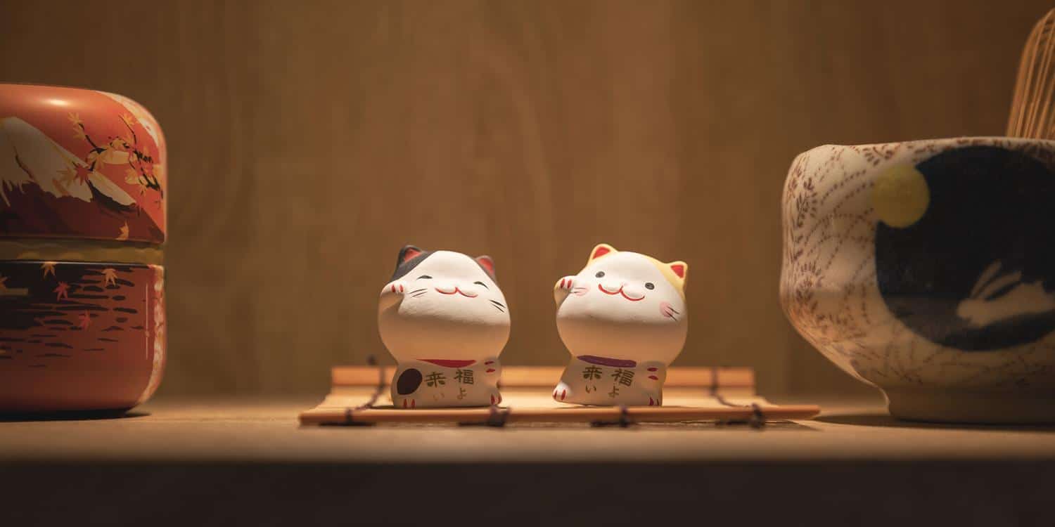 Deux petites figurines "Les chats porte-bonheur" (Taïwan) de Fine Art Shots, souvent connues sous le nom de Maneki Neko ou chats porte-bonheur, sont exposées sur une surface en bois. Un chat est blanc avec une oreille noire et l'autre est blanc avec une oreille jaune. A proximité se trouvent un récipient peint et un bol à motifs.