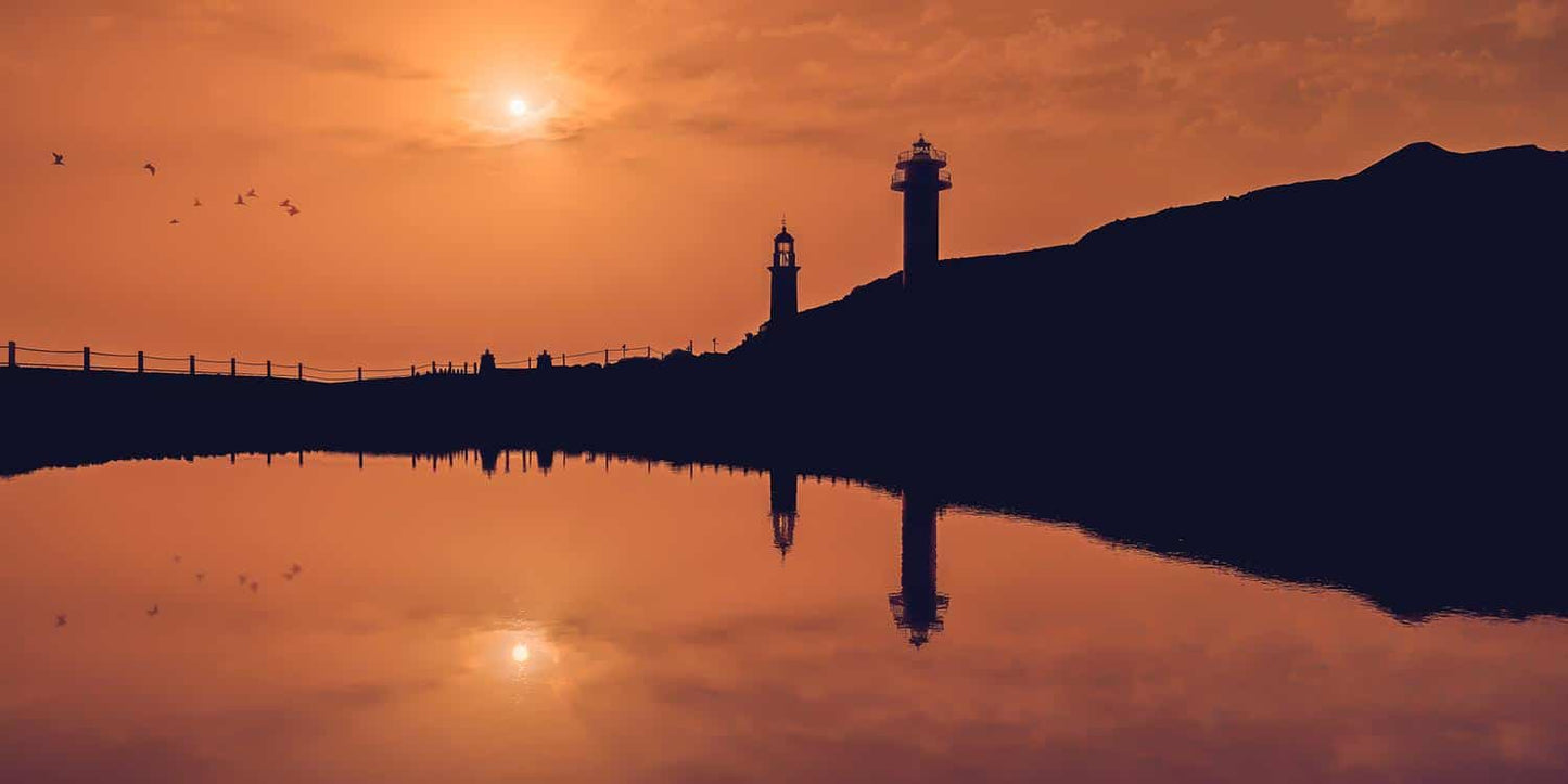 Une scène sereine au coucher du soleil avec deux phares au loin se découpant sur un ciel orange. Les phares et un groupe d'oiseaux volants se reflètent dans l'eau calme en contrebas, avec de légers contours de personnes le long du rivage. "Un mercredi soir sur la terre" (Îles Canaries) de Fine Art Shots capture parfaitement ce moment à couper le souffle.