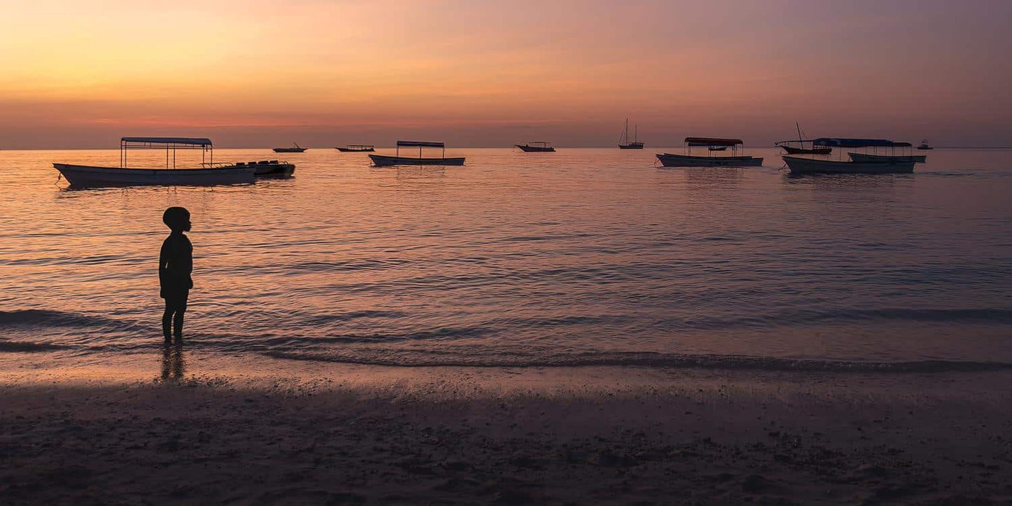 Un enfant se tient sur une plage au coucher du soleil et regarde plusieurs bateaux flotter doucement sur l’eau calme. Le ciel est un mélange d'oranges et de violets chauds, projetant une lueur sereine sur la scène - une image digne des tirages "L'enfant et la plage" (Tanzanie) de Fine Art Shots avec livraison gratuite.