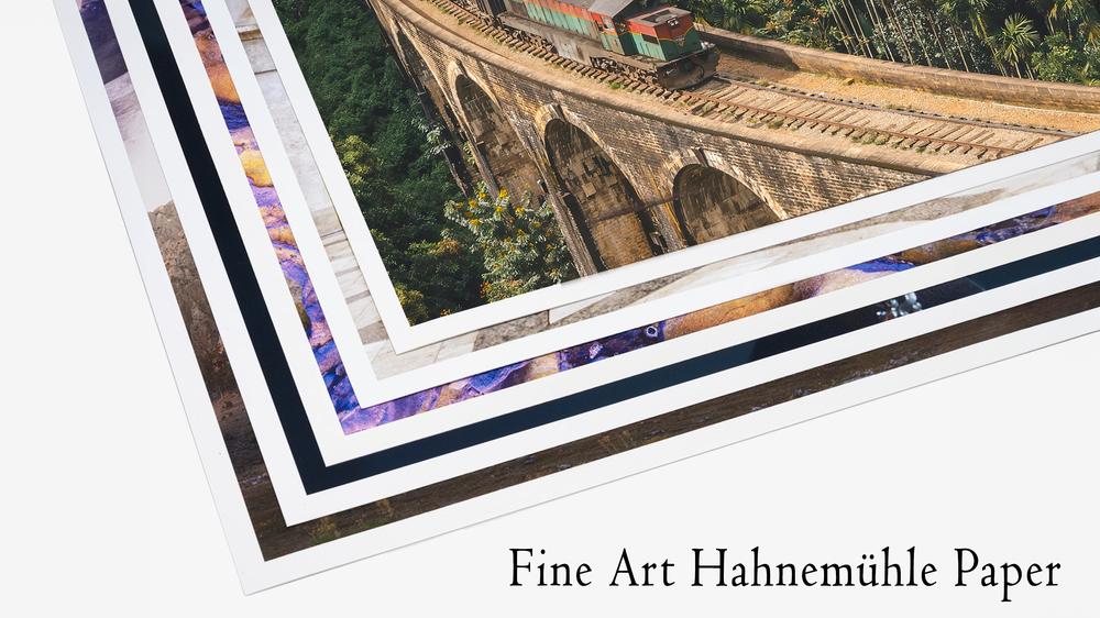 Une pile de tirages de photographies d'art sur papier Hahnemühle est exposée, avec des bords visibles montrant diverses œuvres d'art. La photo du haut montre un train traversant un pont à neuf arches entouré d'une verdure luxuriante. Le texte « Fine Art Hahnemühle Paper » se trouve dans le coin inférieur droit.