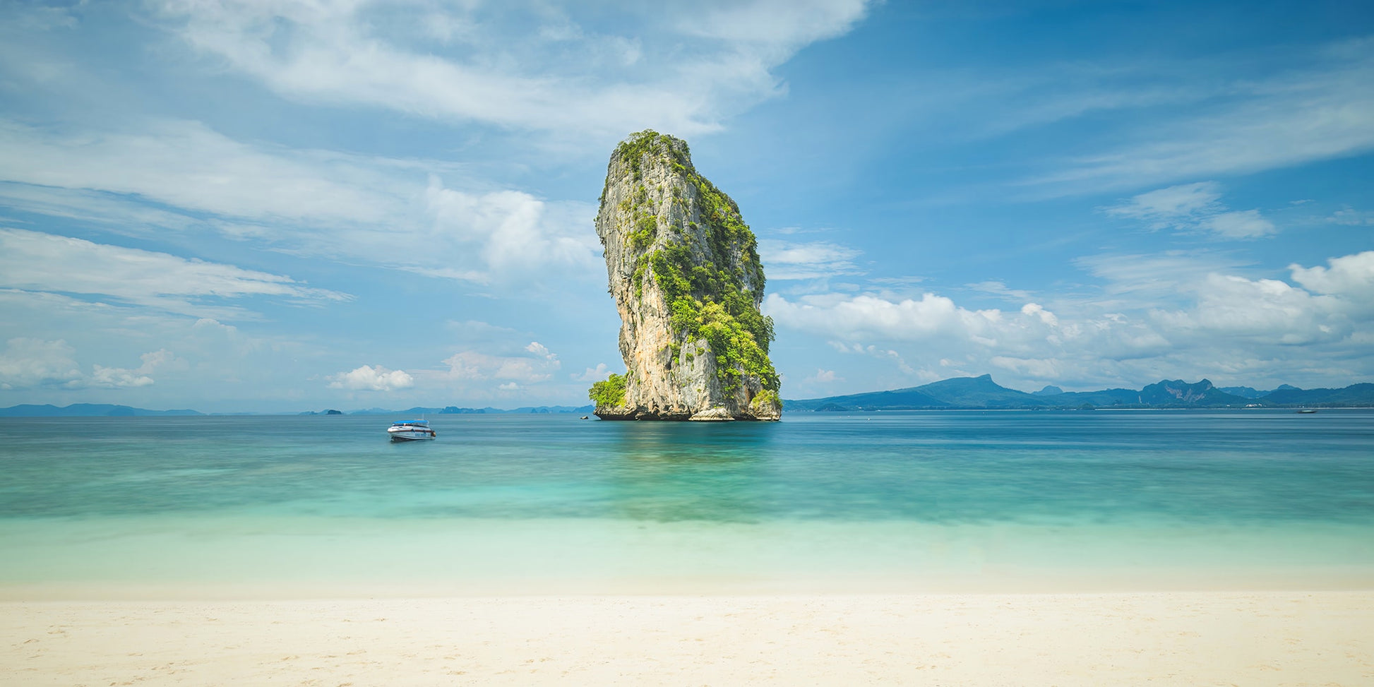 Une scène de plage sereine présente des eaux claires et turquoise et une petite île rocheuse couverte de verdure. Un bateau solitaire est ancré près de l’île sous un ciel bleu vif avec des nuages épars, tandis que des montagnes lointaines sont visibles à l’horizon. "Vue sur rocher" (Thaïlande) de Fine Art Shots capture magnifiquement ce moment tranquille.